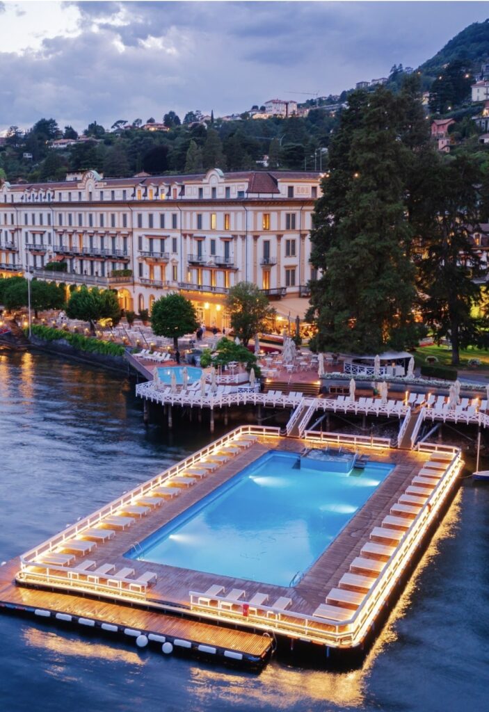 Ein beleuchteter, schwimmender Pool auf einem See neben einem Grand Hotel in der Abenddämmerung, umgeben von Bäumen und Bergen – eine traumhafte Kulisse, perfekt für alle, die nach luxuriösen Locations für eine Hochzeit im Ausland suchen. Diese atemberaubende Kulisse zählt zu den außergewöhnlichsten Hochzeitslocations und ist ideal für eine unvergessliche Hochzeit am Comer See.