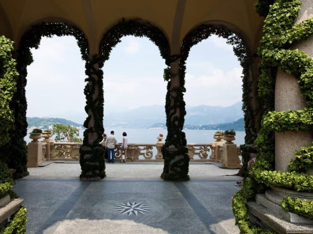 Drei Menschen stehen auf einer Terrasse mit Blick auf das ruhige Wasser des Comer Sees, umrahmt von mit Efeu bewachsenen Bögen und fernen Bergen – die perfekte Kulisse für alle, die nach luxuriösen Hochzeitslocations suchen.