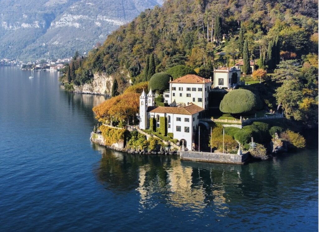 Eine Villa mit roten Dächern am Seeufer, umgeben von Bäumen und Hügeln, die sich im Wasser spiegeln – eine idyllische Kulisse, perfekt für eine luxuriöse Hochzeit an einem besonderen Ort oder eine unvergessliche Hochzeit am Comer See. Diese atemberaubende Villa sticht unter den Hochzeitslocations durch ihre atemberaubende Aussicht und ihr romantisches Ambiente hervor.