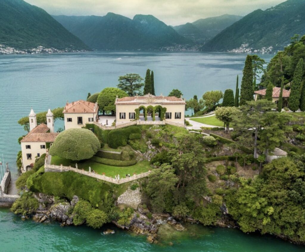 Eine Villa mit Garten auf einem üppig bewachsenen Hügel am Seeufer, umgeben von Bergen und Wasser unter einem bewölkten Himmel – eine idyllische Kulisse für eine Hochzeit am Comer See. Entdecken Sie eine der bezauberndsten Hochzeitslocations, perfekt für Ihre luxuriöse Hochzeit im Ausland.