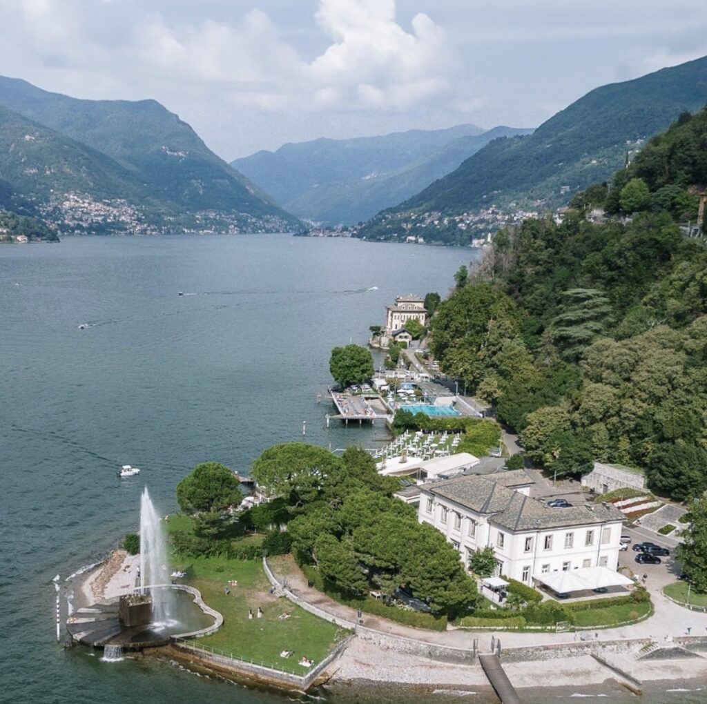 Luftaufnahme einer Stadt am See mit einem Springbrunnen, grünen Hügeln und Bergen im Hintergrund – eine idyllische Kulisse, ideal für eine luxuriöse Hochzeit im Ausland oder für alle, die nach unvergesslichen Hochzeitslocations am Wasser suchen.