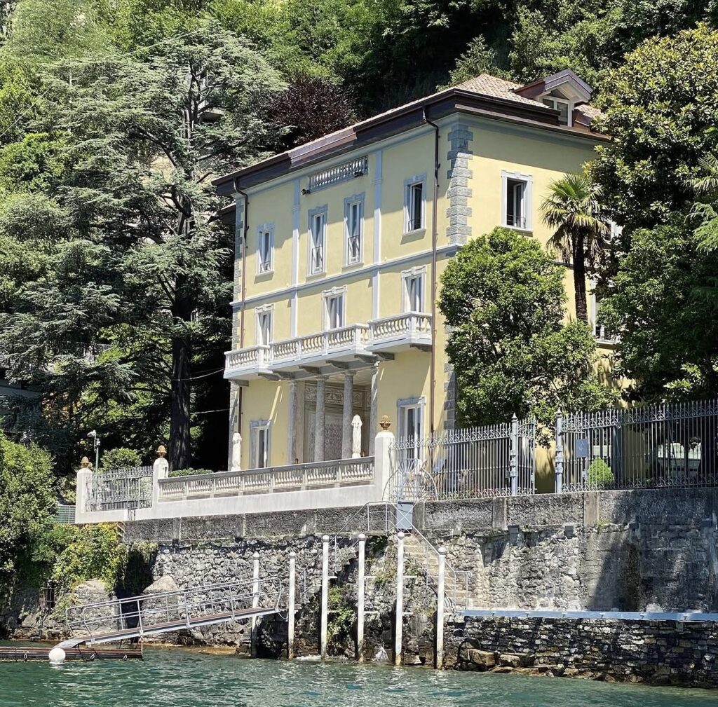 Eine gelbe Villa mit Balkonen und Säulen, umgeben von Bäumen, direkt an einem See mit einem kleinen Steg – ideal für eine luxuriöse Hochzeit im Ausland. Dieses atemberaubende Anwesen ist einer der begehrtesten Hochzeitsorte für eine unvergessliche Hochzeit am Comer See.