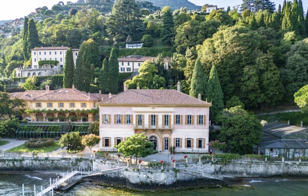 Eine elegante Villa mit hellbraunen Wänden und rotem Dach, umgeben von üppigem Grün direkt am Wasser, mit einem Hügel im Hintergrund – ein idealer Ort für eine luxuriöse Hochzeit am Reiseziel. Dieses exquisite Anwesen ist perfekt für alle, die von einer Hochzeit am Comer See träumen, und bietet eine der bezauberndsten Hochzeitslocations mit atemberaubender Aussicht und zeitloser Eleganz.