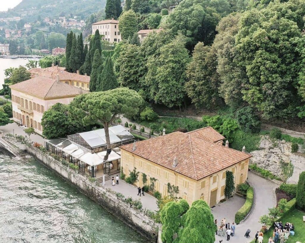 Luftaufnahme einer Villa am Seeufer mit Terrakottadächern, üppigem Baumbestand und Menschen, die am Wasser entlang spazieren – eine bezaubernde Kulisse, ideal für eine luxuriöse Hochzeit am Comer See, einem der begehrtesten Hochzeitsorte Italiens.