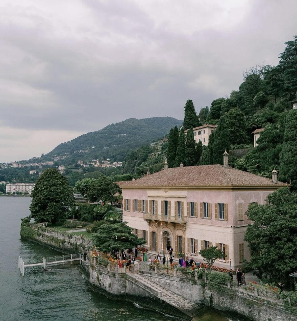 Eine große Villa am See mit Bergen und Bäumen im Hintergrund unter bewölktem Himmel – dieses atemberaubende Anwesen ist einer der begehrtesten Hochzeitsorte für eine luxuriöse Hochzeit am Comer See.