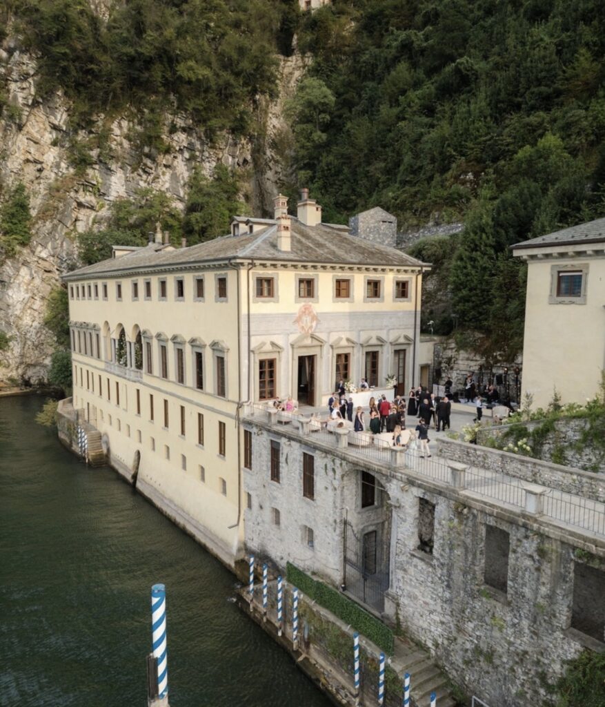 Vor einem großen historischen Gebäude an einer Klippe am Seeufer versammelt sich eine Gruppe von Menschen und schafft so die perfekte Kulisse für alle, die auf der Suche nach exklusiven Hochzeitslocations sind. Die elegante Umgebung versprüht den Charme einer luxuriösen Hochzeit im Ausland und erinnert an eine traumhafte Hochzeitsfeier am Comer See.