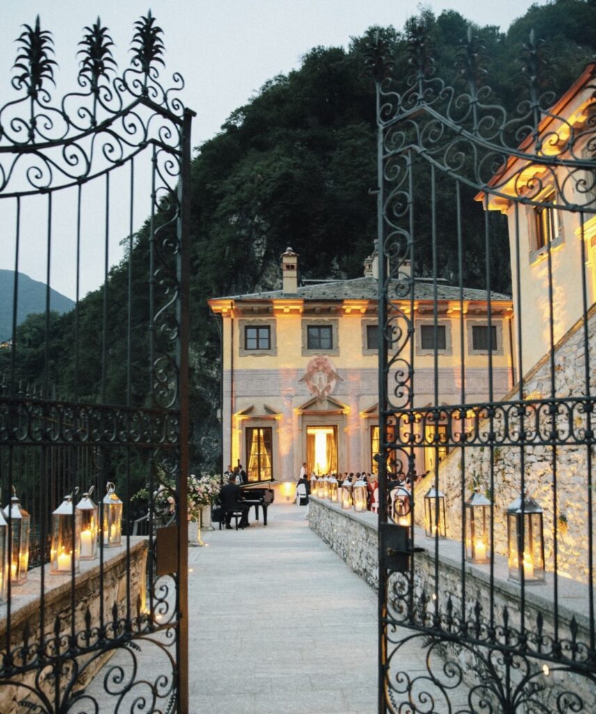Verschnörkelte Eisentore öffnen sich zu einem beleuchteten Steinweg, der in der Abenddämmerung zu einer prächtigen, von Bergen umgebenen Villa führt – eine perfekte Kulisse für eine luxuriöse Hochzeit im Ausland oder eine unvergessliche Hochzeit am Comer See.