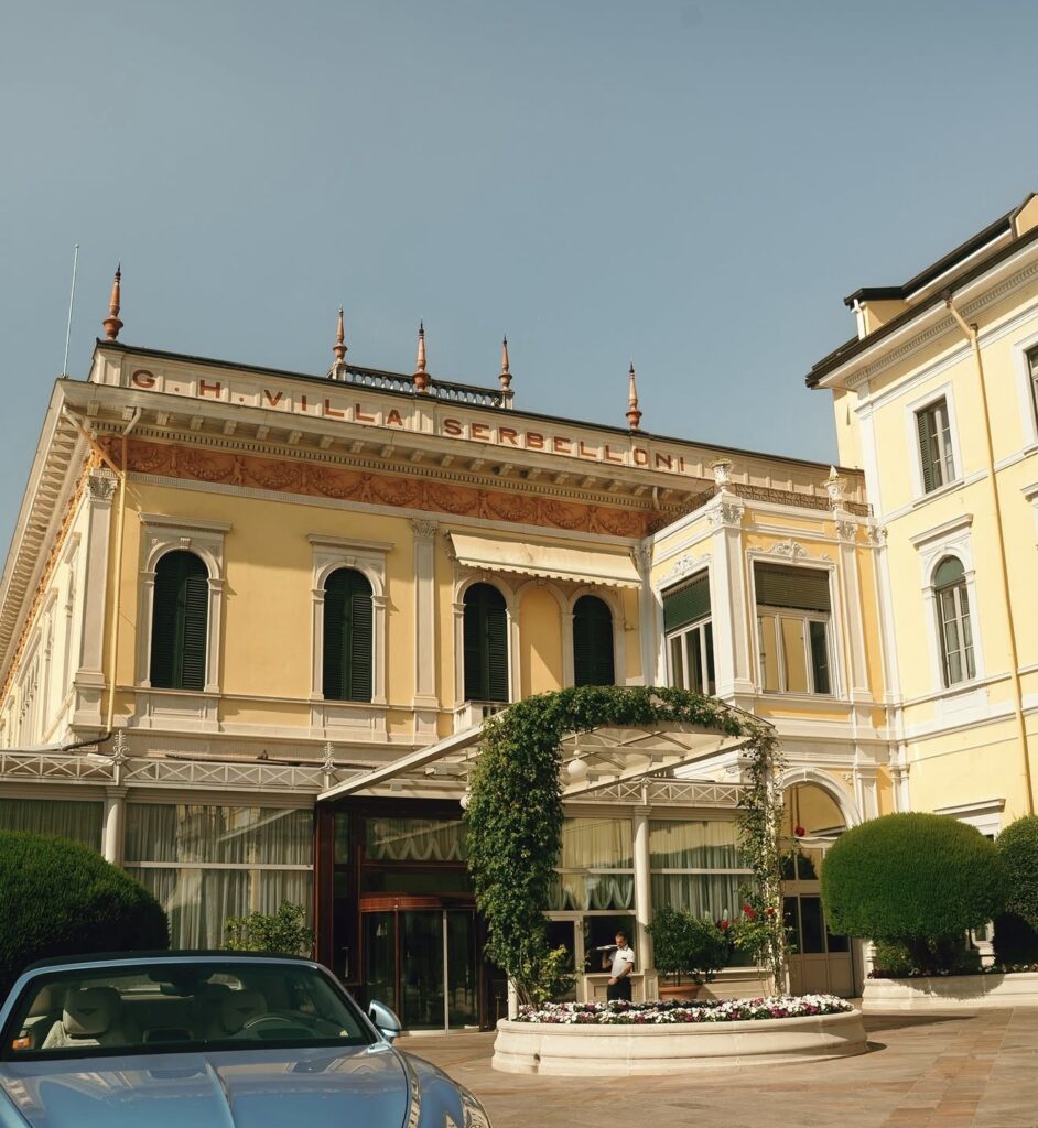 Das Grand Hotel Villa Serbelloni mit seiner eleganten gelben Fassade und den Bogenfenstern gilt als einer der begehrtesten Hochzeitsorte für eine luxuriöse Hochzeit im Ausland. Ein auffälliges blaues Cabrio, das vor dem Hotel geparkt ist, trägt zum Charme bei und bildet die perfekte Kulisse für eine Hochzeit am Comer See.