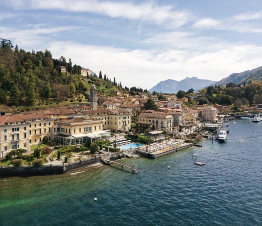 Eine Stadt am See mit Gebäuden direkt am Wasser, einem Steg und grünen Hügeln im Hintergrund unter blauem Himmel – dieser bezaubernde Ort ist perfekt für eine luxuriöse Hochzeit im Ausland. Entdecken Sie eine der begehrtesten Hochzeitslocations, ideal für Ihre Traumhochzeit am Comer See.