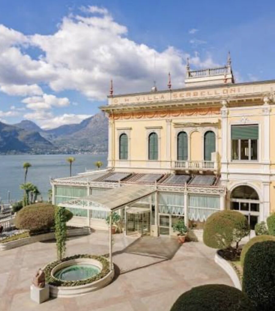 Eine historische Villa am See mit gepflegten Gärten, den Bergen im Hintergrund und einem blauen Himmel mit Wolken – eine idyllische Kulisse für eine luxuriöse Hochzeit an einem besonderen Ort oder eine unvergessliche Hochzeit am Comer See, die zu den exklusivsten Hochzeitslocations der Region zählt.