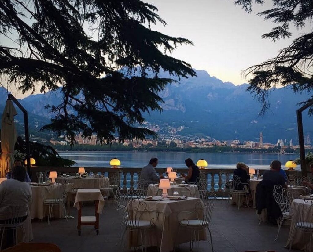 Ein Restaurant im Freien mit Tischen, Gästen beim Essen, einem See und Bergen im Hintergrund bei Sonnenuntergang – eine perfekte Kulisse für eine luxuriöse Hochzeit an einem Traumziel oder eine unvergessliche Hochzeit am Comer See. Diese idyllische Szene zeigt eine der begehrtesten Hochzeitslocations für Paare, die von einer romantischen Feier inmitten atemberaubender Natur träumen.