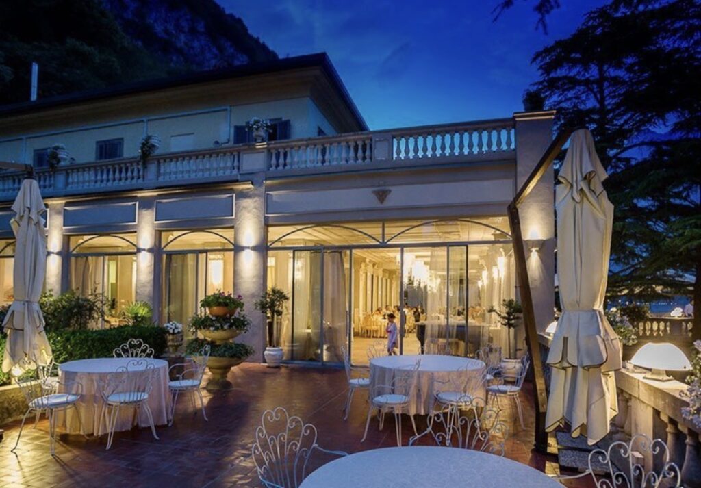 Eine elegante Restaurantterrasse im Freien in der Abenddämmerung mit runden Tischen, weißen Stühlen und leuchtenden Lichtern neben einer Villa – eine idyllische Kulisse, perfekt für eine luxuriöse Hochzeit im Ausland oder für alle, die nach exquisiten Hochzeitslocations für ihre Hochzeit am Comer See suchen.