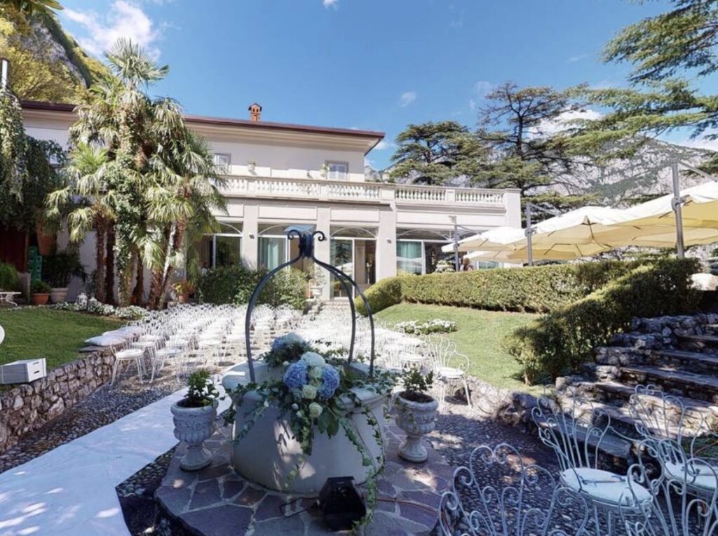 Eine Hochzeitskulisse im Freien mit weißen Stühlen, wunderschönen Blumenarrangements und einer großen Villa im Hintergrund an einem sonnigen Tag – perfekt für alle, die sich eine luxuriöse Hochzeit im Ausland an einer der atemberaubendsten Hochzeitslocations am Comer See wünschen.