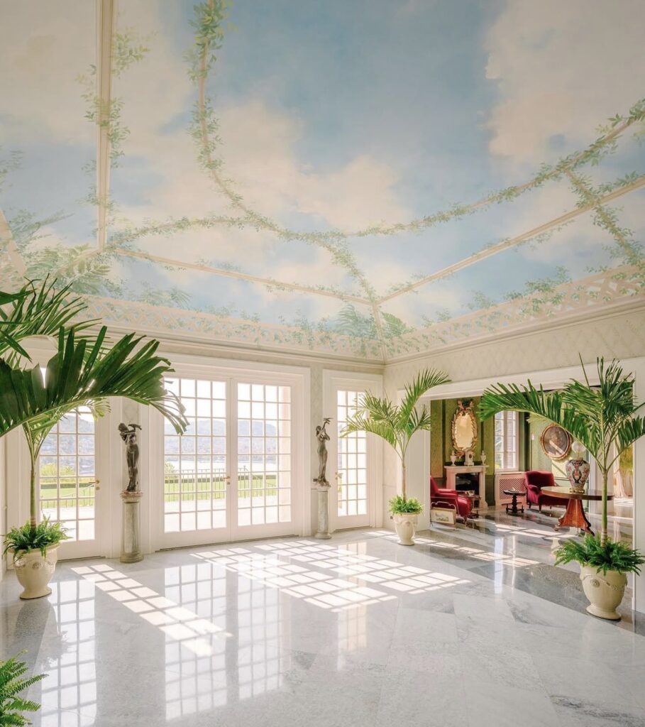 Ein eleganter Wintergarten mit Marmorboden, hohen Topfpalmen, Statuen und einer Deckenmalerei, die den Himmel mit üppigen Ranken darstellt – ein exquisites Ambiente, das an die luxuriösesten Hochzeitslocations erinnert und sich perfekt für eine stilvolle Hochzeit am Comer See oder jede andere luxuriöse Hochzeit im Ausland eignet.