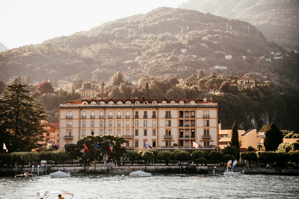 Ein großes Hotel am Seeufer mit Bergen im Hintergrund, Bäumen und kleinen Booten auf dem Wasser davor.