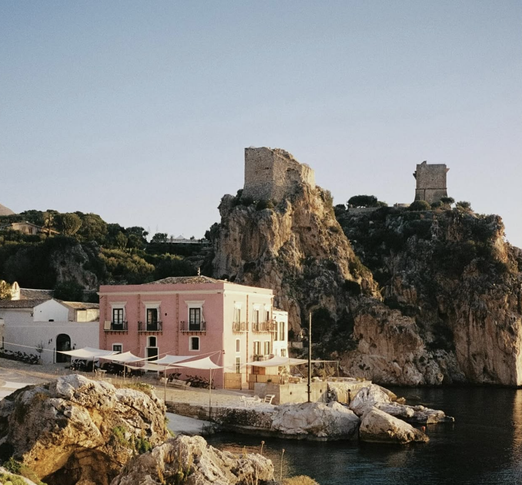 Ein rosa Gebäude am Meer mit felsigen Klippen und antiken Ruinen im Hintergrund unter strahlend blauem Himmel – eine Atmosphäre, die an die bezaubernden Luxus-Hochzeitslocations in Italien wie die Tonnara di Scopello auf Sizilien erinnert.