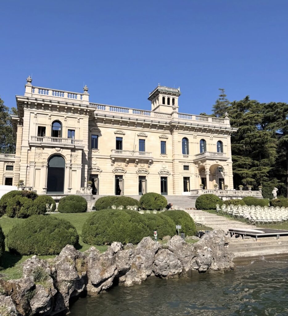 Eine elegante Steinvilla mit Bögen, umgeben von gepflegten Gärten, direkt an einem Gewässer unter strahlend blauem Himmel – sie erinnert an den zeitlosen Charme der Villa Erba am Ufer des Comer Sees.