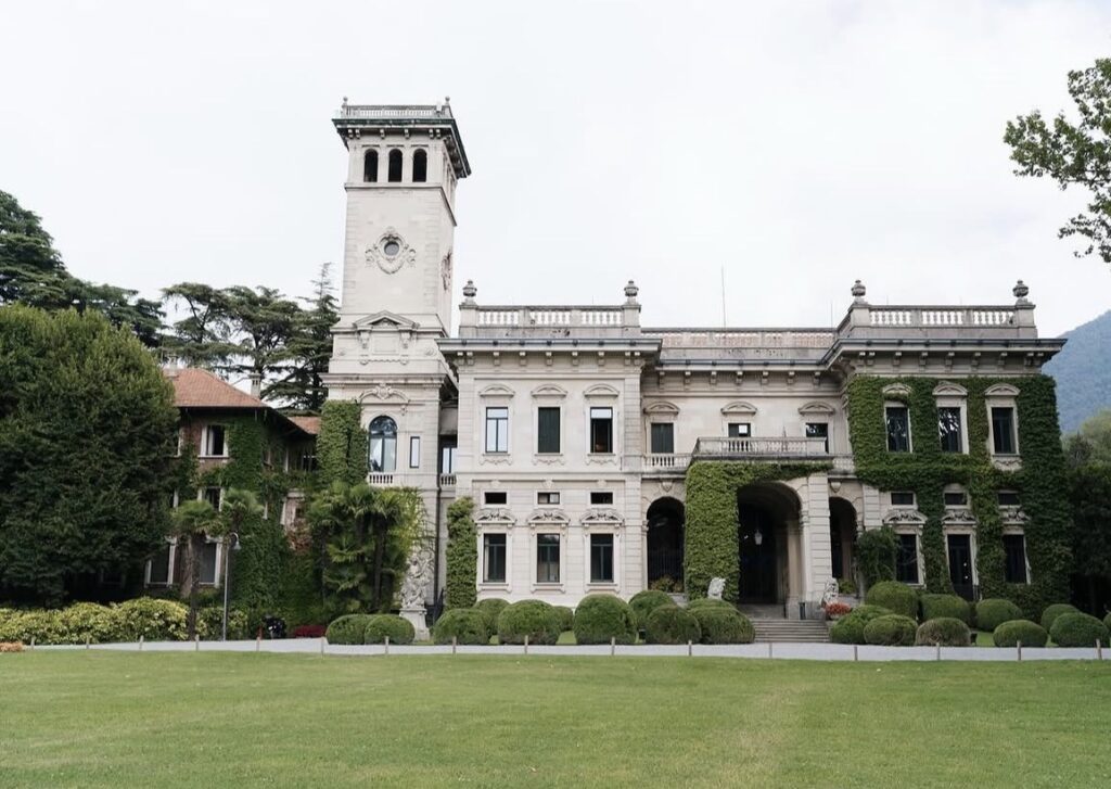 Eine prächtige historische Villa in Erba mit einem hohen Uhrturm, Bogenfenstern und Efeu an den Wänden, umgeben von üppigem Grün am Ufer des atemberaubenden Comer Sees.