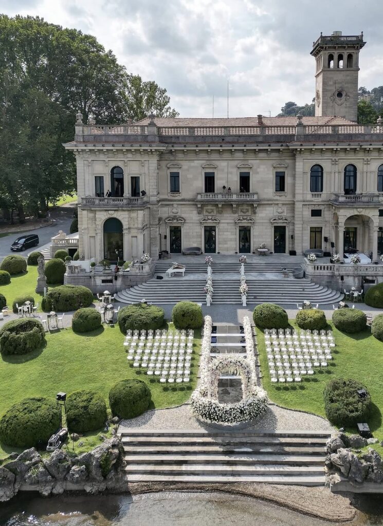 Hochzeitsdekoration im Freien mit weißen Stühlen und eleganten Blumenarrangements vor der prächtigen historischen Villa Erba am Wasser, die einen atemberaubenden Blick auf den Comer See (Lago di Como) bietet.