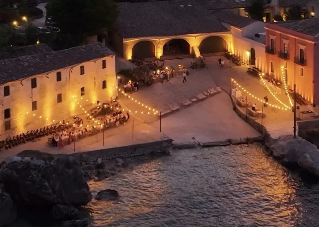 Ein Innenhof am Meer in der Abenddämmerung mit Lichterketten, gedeckten Tischen für das Abendessen und warm beleuchteten Gebäuden – ein Ambiente, das an bezaubernde Hochzeitslocations in Italien wie Borgo Ritella erinnert.