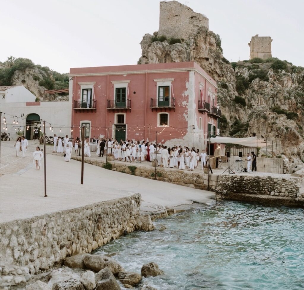 Menschen in weißer Kleidung versammeln sich vor einem rosa Gebäude in der Nähe von türkisfarbenem Wasser, im Hintergrund sind felsige Klippen und antike Ruinen zu sehen – eine bezaubernde Szenerie, die an die Tonnara di Scopello auf Sizilien erinnert, eine der renommiertesten Luxus-Hochzeitslocations in Italien.