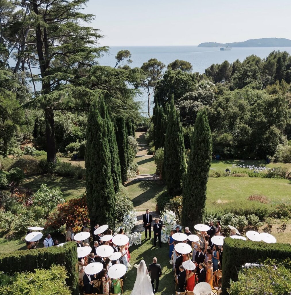 Eine Hochzeitszeremonie in einem üppigen Garten mit Meerblick in der Villa Rocabella, bei der die Gäste weiße Sonnenschirme in den Händen halten – perfekt für alle, die von einer Hochzeit an der Côte d’Azur träumen.