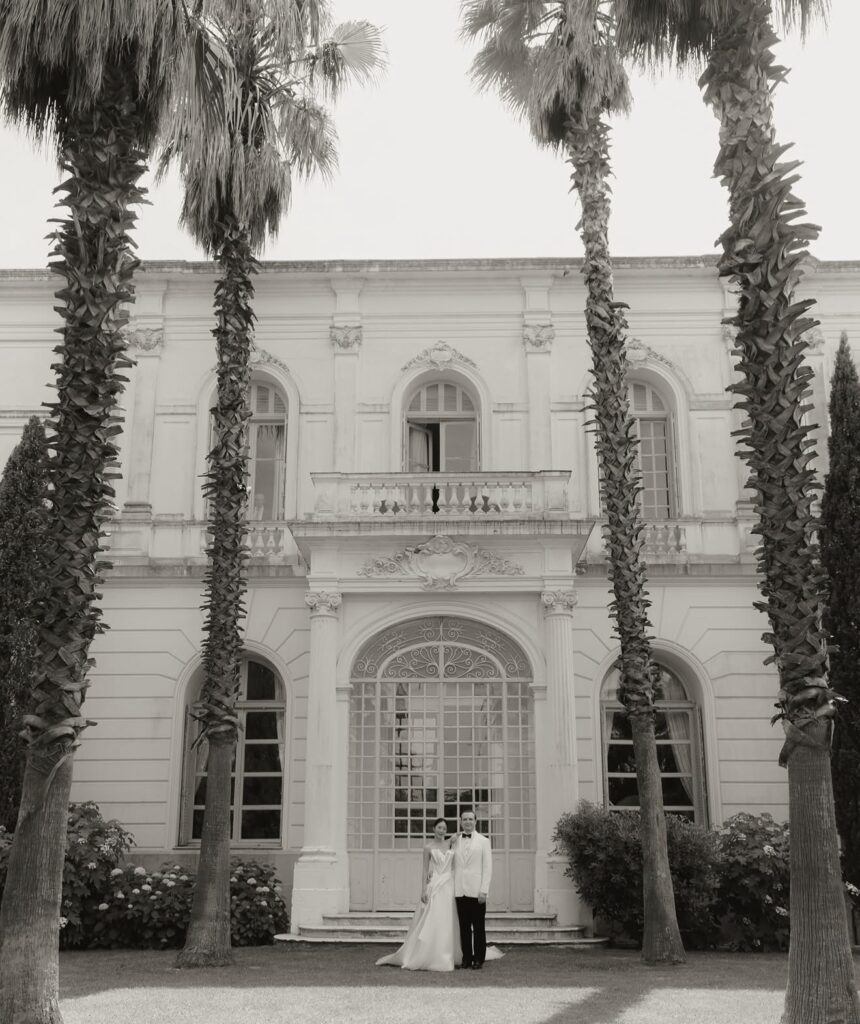 Ein Paar in Hochzeitskleidung steht vor der prächtigen Villa Rocabella, umrahmt von hohen Palmen – eine elegante Kulisse, perfekt für alle, die davon träumen, an der Côte d’Azur zu heiraten.