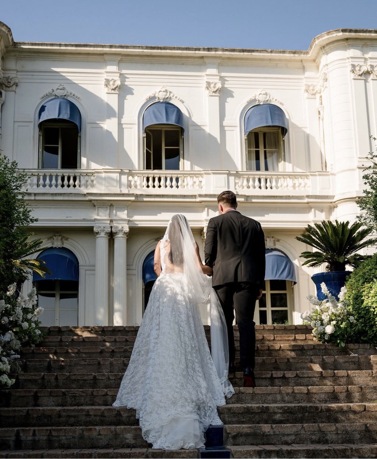 An einem sonnigen Tag schreiten Braut und Bräutigam eine Steintreppe hinauf zu einer weißen Villa mit blauen Markisen – ein Bild, das die romantische Eleganz einer Hochzeit an der Côte d’Azur in der atemberaubenden Villa Rocabella einfängt.