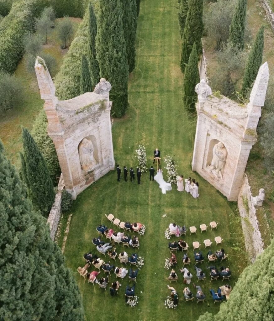 Luftaufnahme einer Hochzeitszeremonie im Freien in einem Garten mit alten Steinbögen und sitzenden Gästen, die einen der schönsten Orte für eine Hochzeit in der Toskana einfängt.