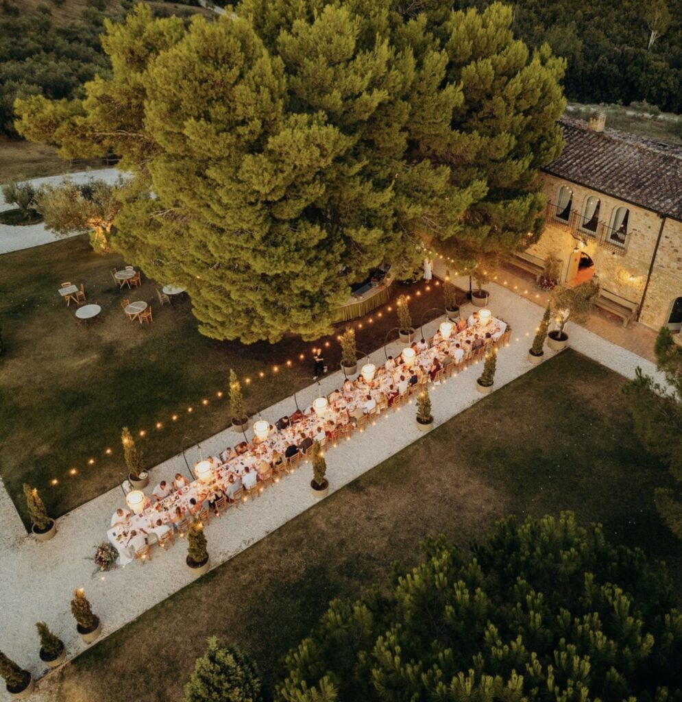 Ein Abendessen im Freien mit einem langen Tisch unter Bäumen im Herzen der Toskana, Gäste, die unter Lichterketten sitzen, und ein rustikales Gebäude in der Nähe – das schafft die perfekte Atmosphäre für eine Hochzeit oder für alle, die auf der Suche nach bezaubernden Hochzeitslocations in der Toskana sind.