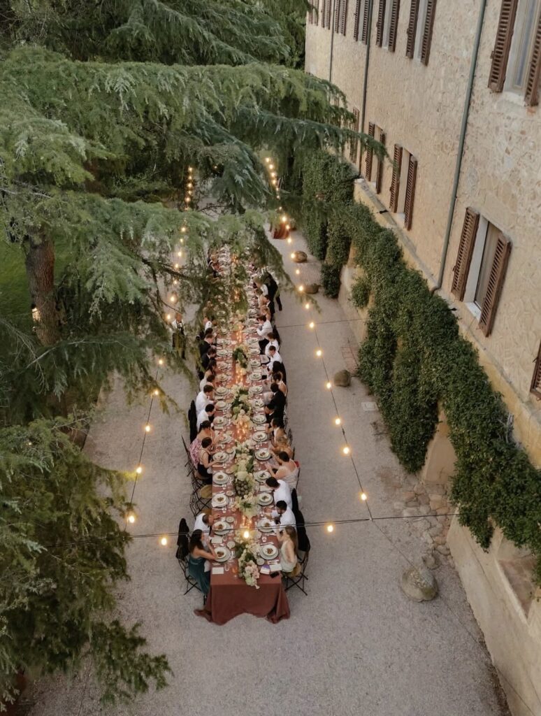 Ein langer Esstisch im Freien unter Lichterketten, umgeben von Menschen, direkt neben einem mit Efeu bewachsenen Steingebäude – einer der schönsten Orte für eine Hochzeit in der Toskana.