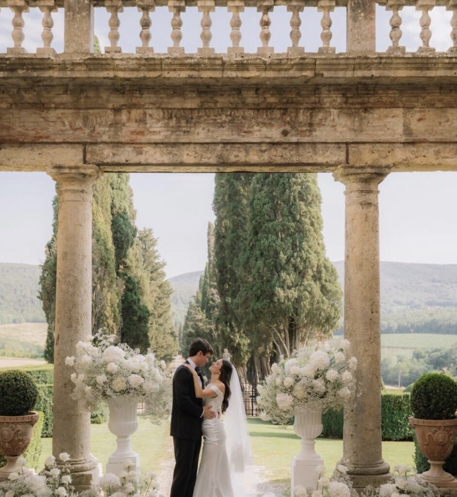 Ein Brautpaar umarmt sich unter einem steinernen Torbogen, umgeben von Blumen und Bäumen in einer romantischen Gartenkulisse in der Toskana – ein perfekter Moment, um ihre Hochzeit zu feiern. Diese bezaubernde Szene fängt die zeitlose Schönheit der Hochzeitslocations in der Toskana ein, wo die Liebe inmitten der Eleganz der Natur erblüht.