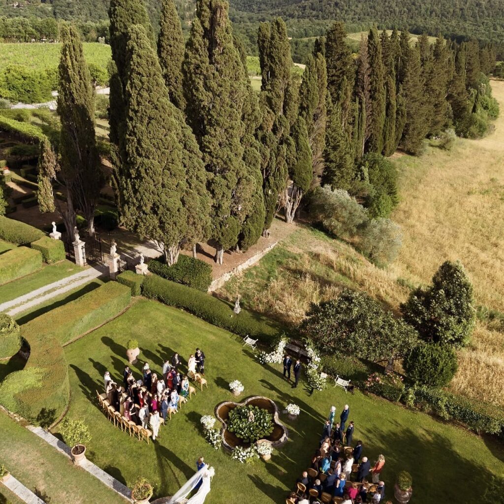Luftaufnahme einer Hochzeitszeremonie im Freien auf einer grünen Wiese, umgeben von Bäumen und der atemberaubenden Landschaft, die den romantischen Charme einer der schönsten Hochzeitslocations in der Toskana einfängt.