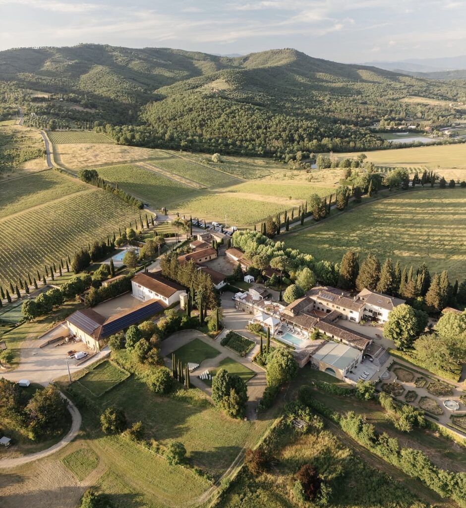 Luftaufnahme eines Landguts, eines der schönsten Orte für unvergessliche Hochzeitslocations in der Toskana, mit Feldern, Bäumen und Hügeln im Hintergrund unter einem teilweise bewölkten Himmel.