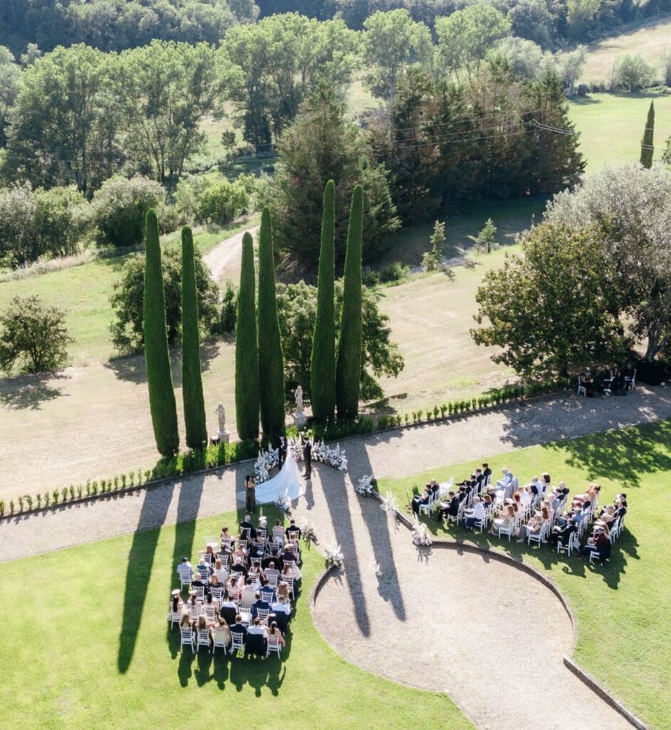 Eine Hochzeitszeremonie im Freien mit in Reihen sitzenden Gästen, hohen Bäumen und üppigem Grün im Hintergrund – einer der schönsten Orte für eine Hochzeit in der Toskana, perfekt für alle, die nach idyllischen Hochzeitslocations in der Toskana suchen.