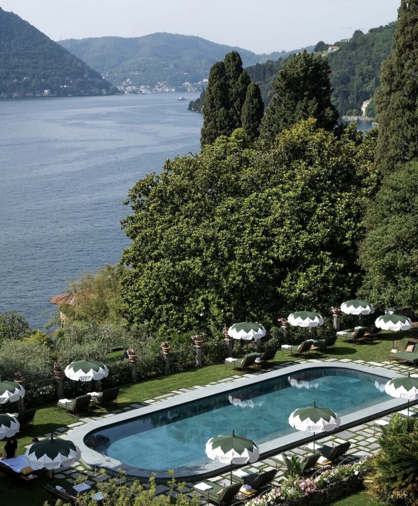 A pool with sunshades in Passalacqua offers a view of Lake Como, surrounded by trees and hills under a clear sky.