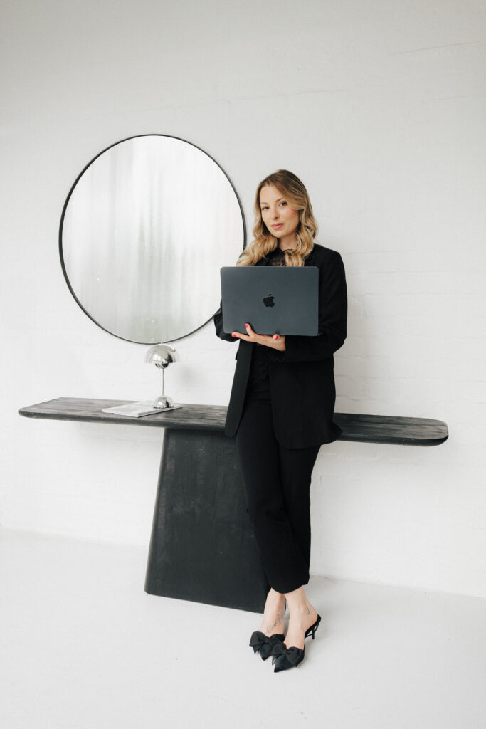 Eine Frau in einem schwarzen Anzug hält einen Laptop in der Hand und lehnt an einem schwarzen Tisch, an dessen Wand ein runder Spiegel hängt – ein Bild, das die moderne Eleganz verkörpert, die perfekt zu jeder VIP-Hochzeit passt.
