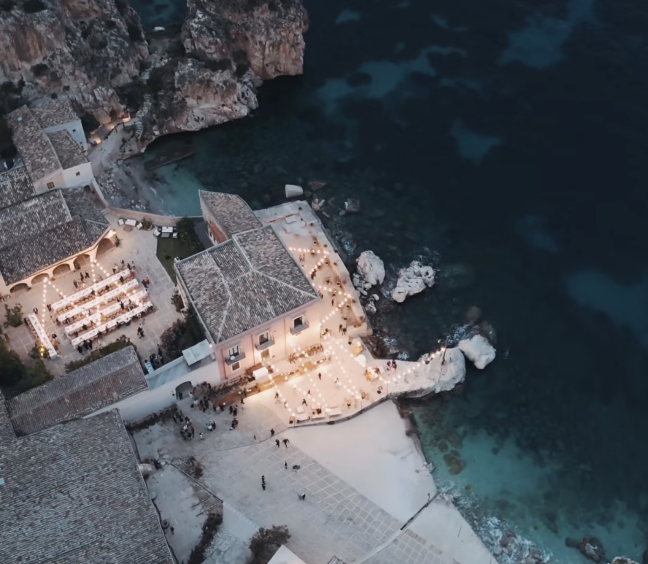 Luftaufnahme eines Restaurants am Meer in der Abenddämmerung mit Außenbeleuchtung und türkisfarbenem Wasser in der Nähe, die an das bezaubernde Ambiente renommierter Luxus-Hochzeitslocations in Italien wie beispielsweise der Tonnara di Scopello erinnert.