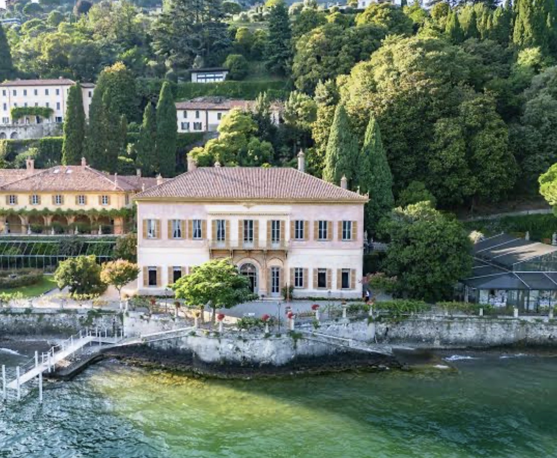 Große Villa mit Terrakottadach an einem See, umgeben von üppigem Grün und hohen Bäumen.