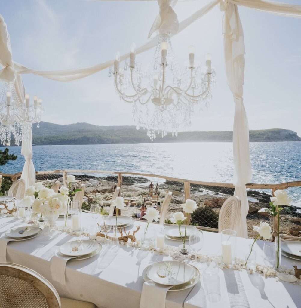 A long dining table, set for a luxurious wedding at the destination on a terrace by the sea under chandeliers.