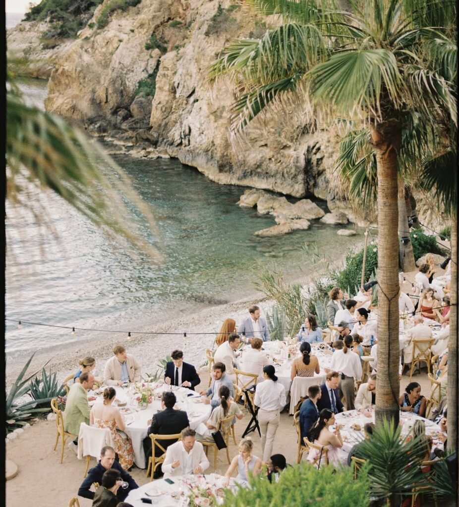 At a luxurious wedding in Ibiza, the guests dine at outdoor tables on the beach near a rocky cliff.