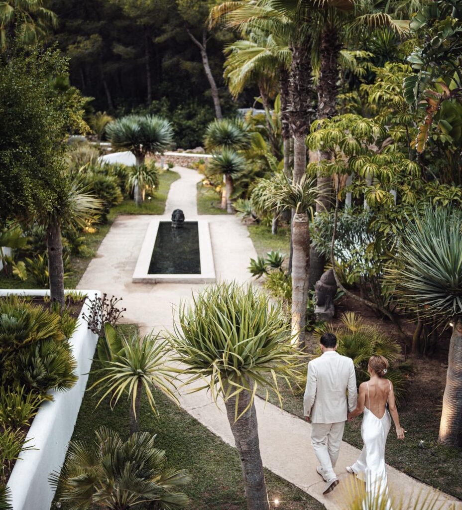 A couple holds hands as they walk through a lush tropical garden at a luxurious wedding venue in Ibiza.